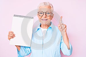 Senior beautiful woman with blue eyes and grey hair holding notebook surprised with an idea or question pointing finger with happy