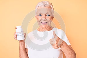 Senior beautiful woman with blue eyes and grey hair holding pills smiling happy and positive, thumb up doing excellent and