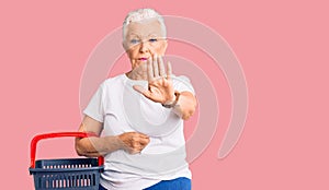 Senior beautiful woman with blue eyes and grey hair holding supermarket shopping basket with open hand doing stop sign with