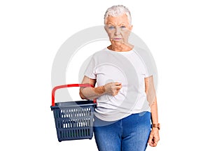 Senior beautiful woman with blue eyes and grey hair holding supermarket shopping basket thinking attitude and sober expression