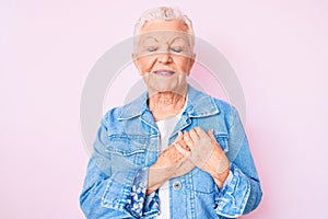 Senior beautiful woman with blue eyes and grey hair wearing casual denim jacket smiling with hands on chest with closed eyes and