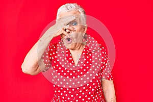 Senior beautiful woman with blue eyes and grey hair wearing casual summer clothes over red background peeking in shock covering