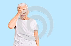 Senior beautiful woman with blue eyes and grey hair wearing casual white tshirt covering eyes with hand, looking serious and sad