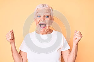 Senior beautiful woman with blue eyes and grey hair wearing classic white tshirt over yellow background celebrating surprised and