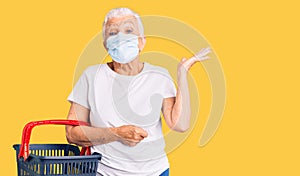 Senior beautiful woman with blue eyes and grey hair wearing shopping basket and medical mask celebrating victory with happy smile