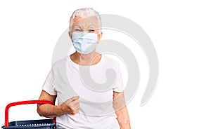 Senior beautiful woman with blue eyes and grey hair wearing shopping basket and medical mask looking positive and happy standing