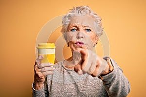 Senior beautiful woman drinking cup of coffee standing over isolated yellow background pointing with finger to the camera and to