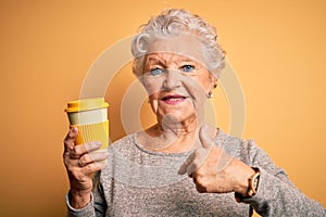Senior beautiful woman drinking cup of coffee standing over isolated yellow background with surprise face pointing finger to