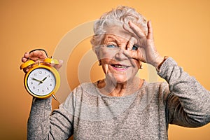 Senior beautiful woman holding alarm clock standing over isolated yellow background with happy face smiling doing ok sign with