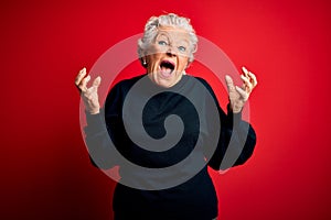 Senior beautiful woman wearing casual sweater standing over isolated red background crazy and mad shouting and yelling with