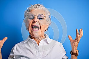 Senior beautiful woman wearing elegant shirt and glasses over isolated blue background crazy and mad shouting and yelling with