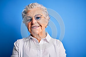 Senior beautiful woman wearing elegant shirt and glasses over isolated blue background happy face smiling with crossed arms