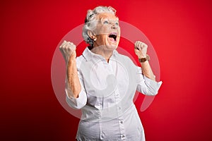 Senior beautiful woman wearing elegant shirt standing over isolated red background crazy and mad shouting and yelling with