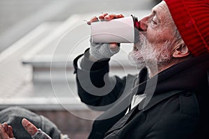 Old-aged vagabond sitting on walking street and drinking coffee or tea photo
