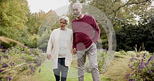 Senior biracial couple holding hands and walking together in sunny garden, unaltered, in slow motion