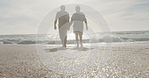 Senior biracial couple walks hand in hand on a sunny beach, with copy space