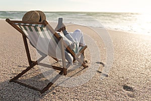 Senior biracial woman sitting on chair using mobile phone while relaxing at beach during sunset