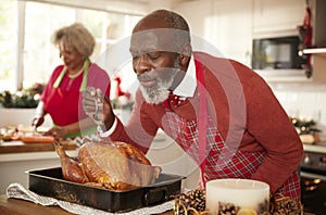 Senior black man basting roast turkey in preparation for Christmas dinner, his wife chopping vegetables in the background, close u