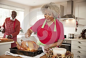 Senior black woman basting a roast turkey in preparation for Christmas dinner, her husband chopping vegetables in the background,