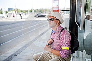 Senior blind man with white cane waiting for public transport in city.