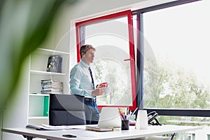 Senior business man holding a coffee cup and taking a phone call on his smart phone whilst standing in his office