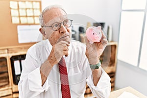 Senior business man holding piggy bank serious face thinking about question with hand on chin, thoughtful about confusing idea