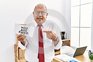 Senior business man holding you are fired banner at the office smiling happy pointing with hand and finger