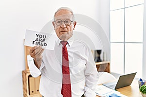 Senior business man holding you are fired banner at the office thinking attitude and sober expression looking self confident