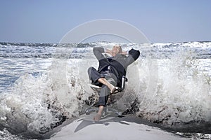 Senior business man sitting on office chair on beach being splashed by waves