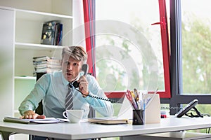 Senior business man working whilst making telephone call, he looks shocked and slamming his hand on his desk