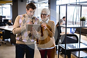 Senior business woman and her young colleague standing in office with digital tablet