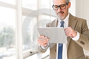 A senior businessman in a beige suit stands by a bright window, smiling as he engages with a tablet