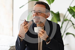 Senior businessman in glasses using voice commands on his smartphone while enjoying an espresso