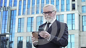 Senior businessman scrolling smartphone app holding coffee cup, lunchtime break