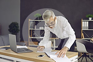 Senior businessman standing by the desk in his office and taking notes in his notebook