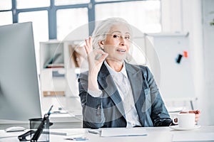 Senior businesswoman drinking coffee and showing ok sign at workplace