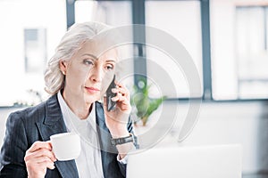 Senior businesswoman holding coffee cup and talking on smartphone