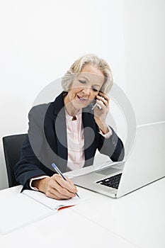 Senior businesswoman writing in book while answering cell phone at desk in office