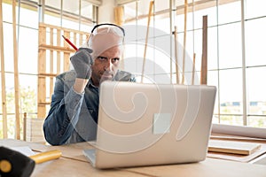 A senior carpenter man wearing glove and goggle looking at laptop screen in the modern wood workplace. Looking the drawing and