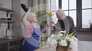 Senior Caucasian blond woman trying to beat her husband with frying pan. Mature baldheaded man defending himself with a