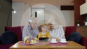 Senior Caucasian couple with cute child girl granddaughter using tablet, watching funny videos