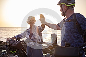 Senior caucasian couple riding off road on the pebble beach with bicycles at sunset giving high five smiling. Authentic elderly