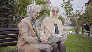 Senior Caucasian female friends in headscarves and sunglasses sitting on the bench and examining the map. Mature elegant