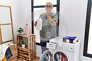 Senior caucasian man doing laundry holding detergent bottle covering mouth with hand, shocked and afraid for mistake