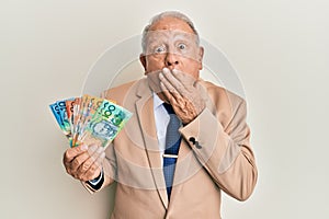 Senior caucasian man holding australian dollars covering mouth with hand, shocked and afraid for mistake