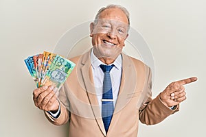 Senior caucasian man holding australian dollars smiling happy pointing with hand and finger to the side