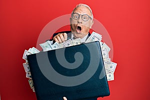 Senior caucasian man holding briefcase full of dollars afraid and shocked with surprise and amazed expression, fear and excited