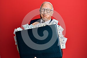 Senior caucasian man holding briefcase full of dollars puffing cheeks with funny face