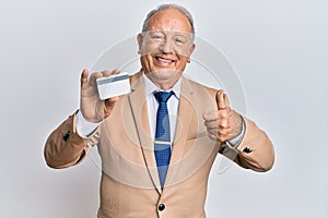 Senior caucasian man holding credit card smiling happy and positive, thumb up doing excellent and approval sign