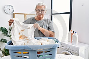 Senior caucasian man holding dirty t shirt with stain winking looking at the camera with sexy expression, cheerful and happy face